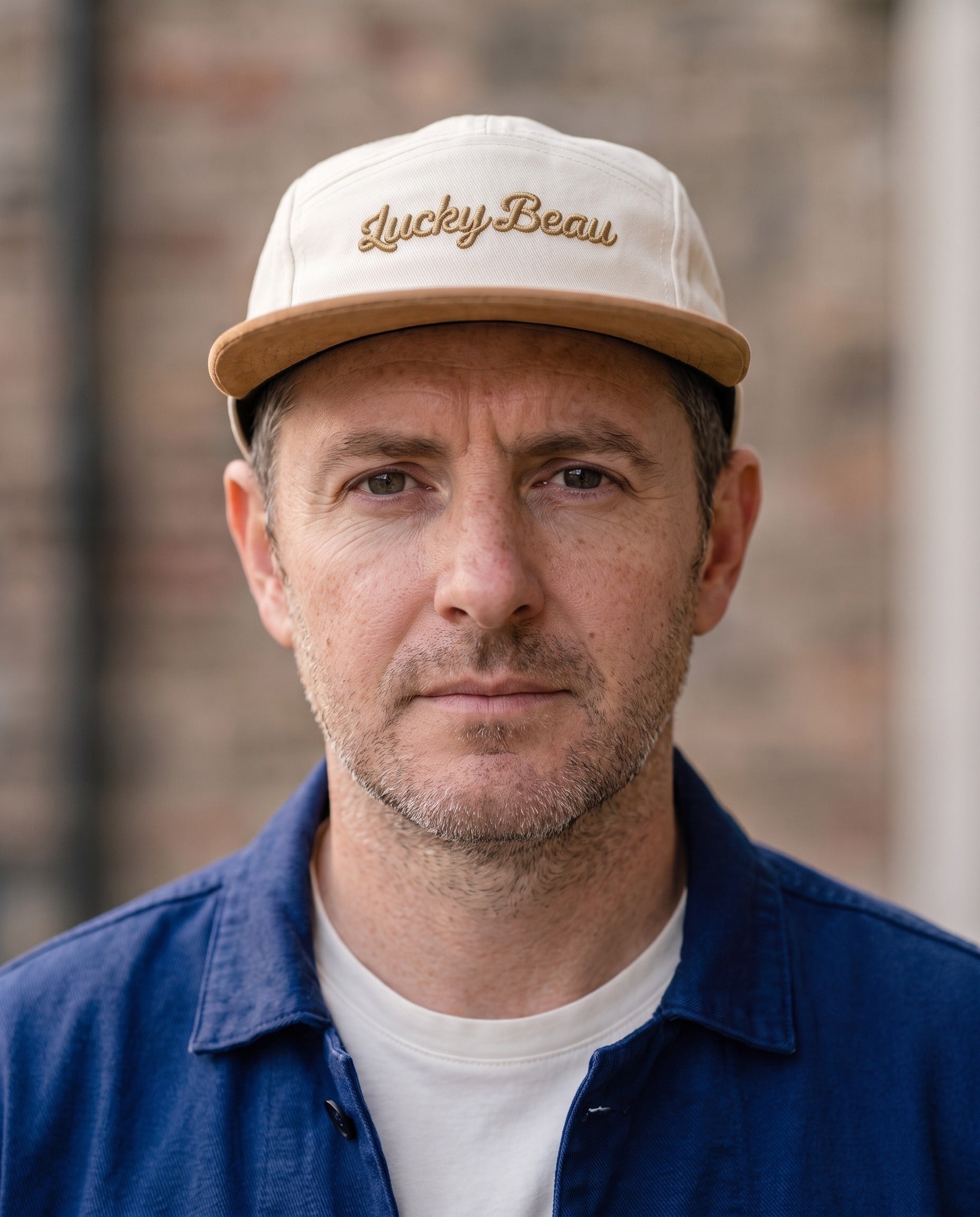 Man wearing the Lucky Beau 5-Panel Cap against a brick wall, paired with the Chasseur Bleu jacket